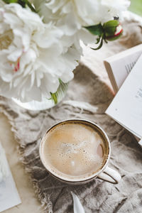 Stylish ceramic cup with  coffee and foam at window with curtains and glass vase peonies, cozy home