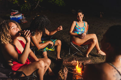 Group of cheerful ethnic friends with beer having fun while gathering around burning bonfire during summer hangout in evening time on beach in costa rica