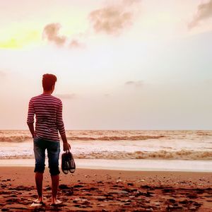 Rear view of man standing on shore at beach during sunset