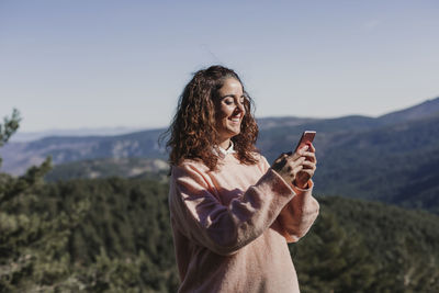 Smiling woman using phone while standing on mountain against sky