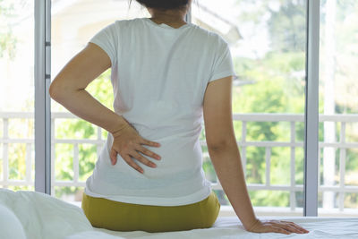 Midsection rear view of woman with backache sitting on bed at home