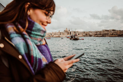 Smiling young woman using mobile phone by sea against sky
