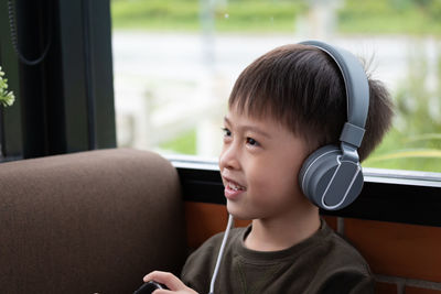 Cute smiling boy looking away while listening music on headphones against window