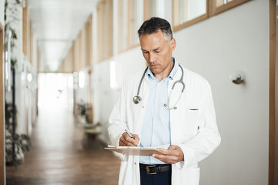 Mature doctor writing medical record at corridor in hospital