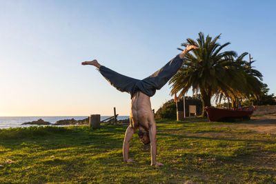 Shirtless and barefoot man standing on hands with spreading and stretching legs on green lonely seaside with exotic palms and blue sky on background