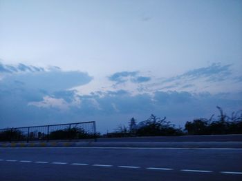 Empty road by silhouette trees against sky
