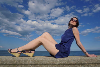 Confident woman in sunglasses sitting on retaining wall against sky
