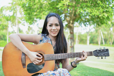 Portrait of smiling young woman playing guitar while sitting at park