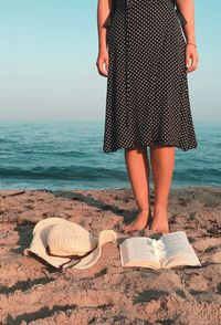 Low section of woman standing by open book and hat at beach against sea