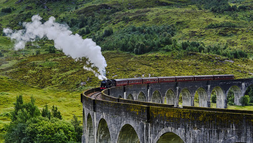 View of steam train crossing arch railway bridge amidst trees and mountains 