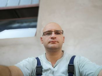 Low angle portrait of bald man wearing eyeglasses while sitting against wall