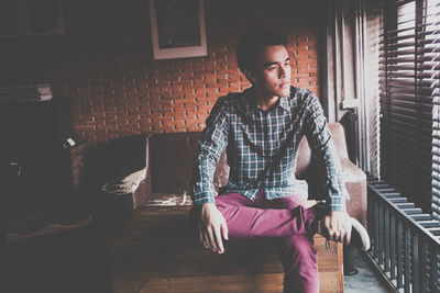 Thoughtful young man looking away while sitting by window blinds at home