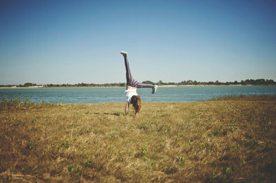 Full length of girl practicing handstand on grassy field by lake against clear sky