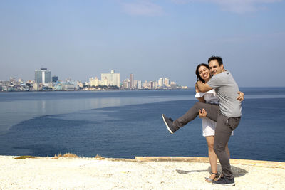 Full length of young woman standing by sea against cityscape