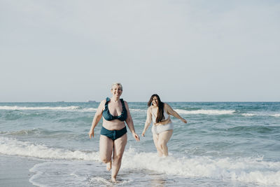 Positive curvy female friends in swimwear walking in sea water during summer vacation looking at camera