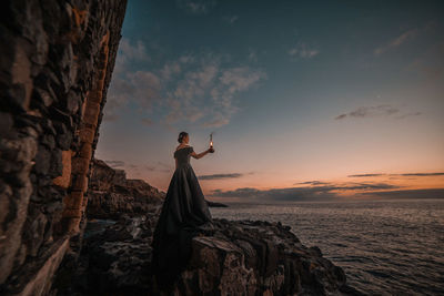 Side view of woman standing on rocky shore against sky during sunset