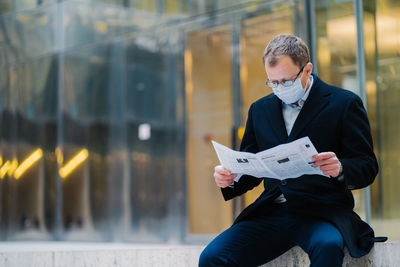 Businessman wearing mask reading newspaper while sitting outdoors