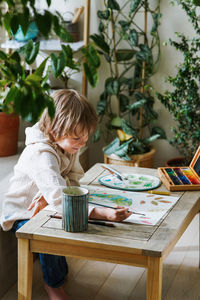 Cute male kid art drawing multicolored image use paints and brush at brightly room with potted plant
