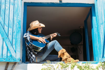 Side view of cheerful ethnic female dressed in cowboy style clothes with boots and hat sitting on windowsill and playing guitar while resting in rural house in summer day