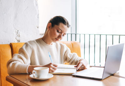 Attractive young smiling woman student freelancer with cup of tea using laptop working in cafe