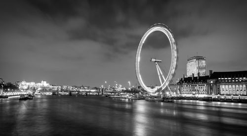 Illuminated ferris wheel by river against sky in city at night