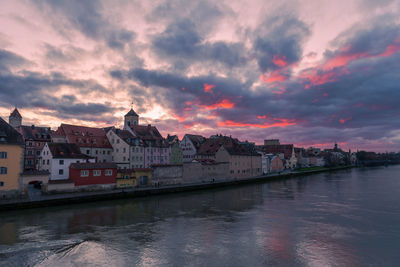 Buildings by river against sky at sunset
