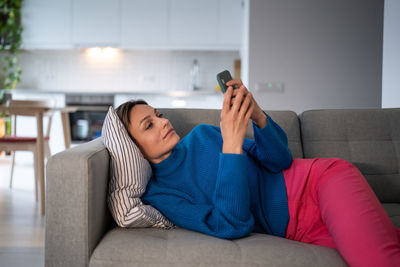 Calm woman in blue sweater rests on comfortable sofa and scrolls social media in internet
