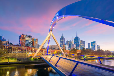 Illuminated bridge over river by buildings against sky in city