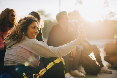 Happy woman taking selfie on smart phone while sitting with friends at park