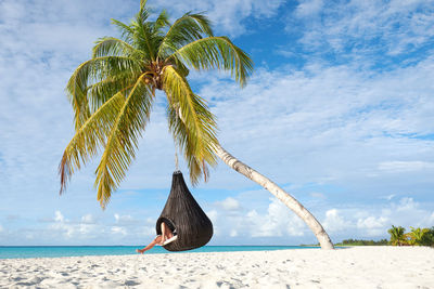 Woman swinging from palm tree on beach against sky