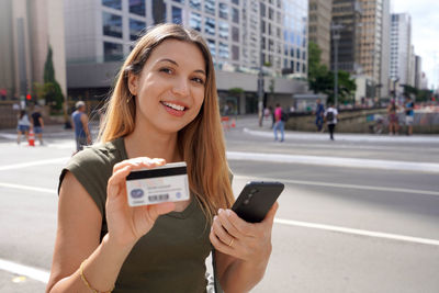 Portrait of smiling woman holding a smartphone and showing a credit card, paulista avenue