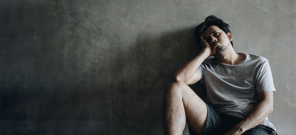 Young man looking away while sitting against wall