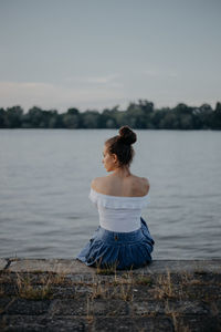 Rear view of woman sitting by lake on retaining wall against sky