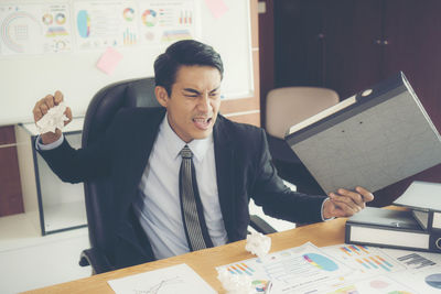 Frustrated businessman holding file while sitting at desk in office