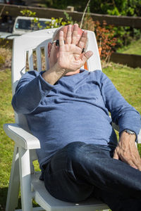 Senior man covering face while sitting on chair during sunny day