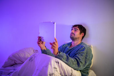 Man using digital tablet sitting on bed against wall at home