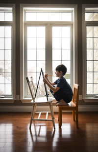 Young boy drawing on a canvas on an easel in front of windows.