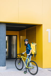 Smiling young delivery woman with bicycle standing in front of building