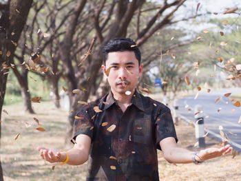 Portrait of young man throwing leaves and twigs while standing against trees