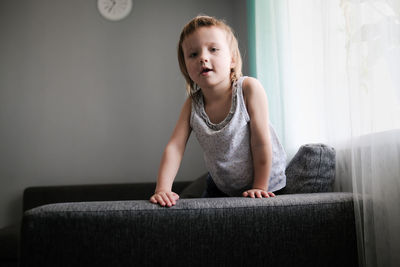 European toddler child sits and gets bored on gray sofa in bright living room, minimalist interior