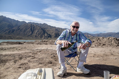 Man smiling while pouring wine in a plastic glass outdoors in natural landscape.