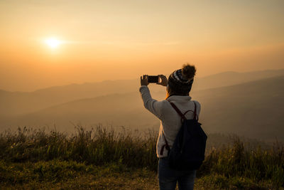 Rear view of woman photographing on mountain against sky during sunset