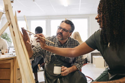 Male tutor assisting female student in painting on artist's canvas in art class