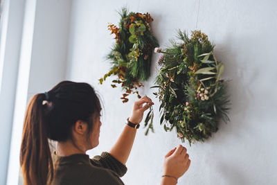 Young woman decorating christmas wreath on wall at home 