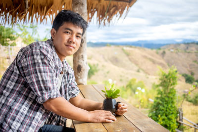 Side view portrait of smiling young man holding plant in cup at hut