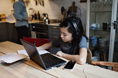 Serious boy doing homework while using laptop and mobile phone at home