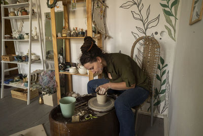 Woman making clay pot on pottery wheel sitting on chair at workshop