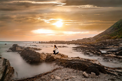 Teenage girl sitting on rock at beach against sky during sunset