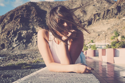Young woman with tousled hair leaning on retaining wall against mountain