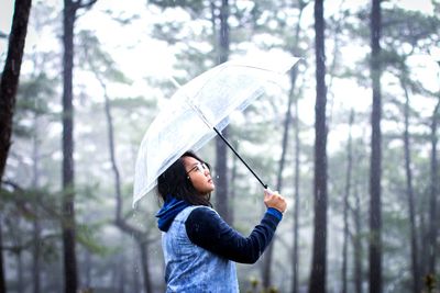 Side view of woman holding umbrella while standing in forest during rainy season